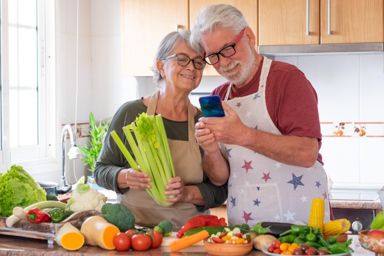 Vegetarian lifestyle. Beautiful elderly couple white-haired enjoying cooking together. Looking at smartphone. On the table a mix the of raw seasonal vegetables